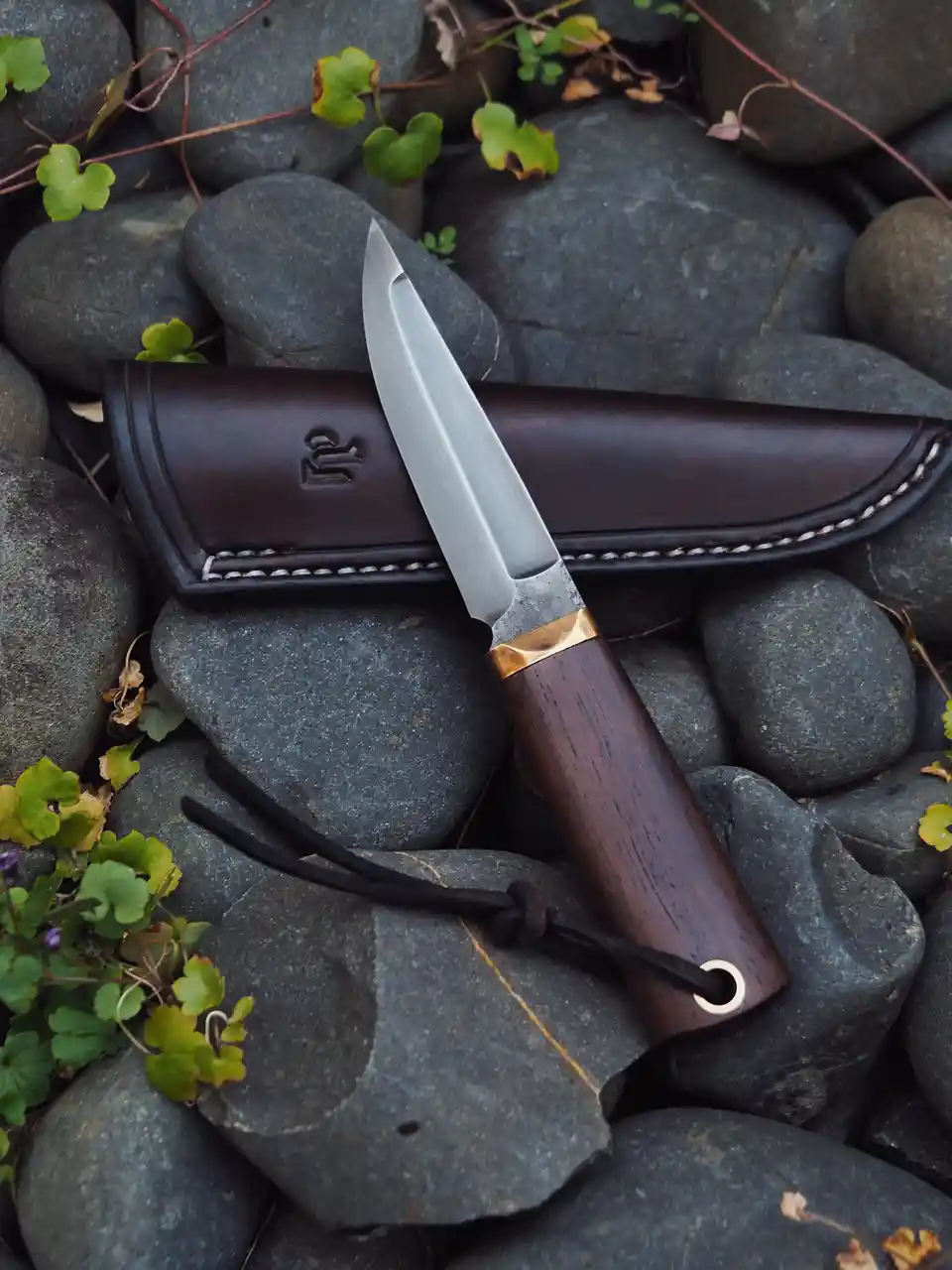 A small, handcrafted knife with a wooden handle and brass bolster rests partially drawn from its dark brown leather sheath…