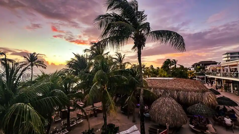 A scenic tropical beach sunset with palm trees, thatched-roof huts, and people relaxing at an outdoor bar. The overlaid text…