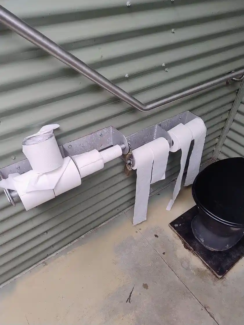 A photo shows multiple toilet paper rolls mounted on a metal bar attached to a corrugated metal wall, with some rolls…