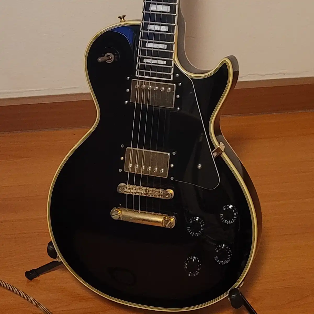 A black electric guitar with gold hardware and binding is propped up on a stand on a wooden floor, with a plain wall in the…