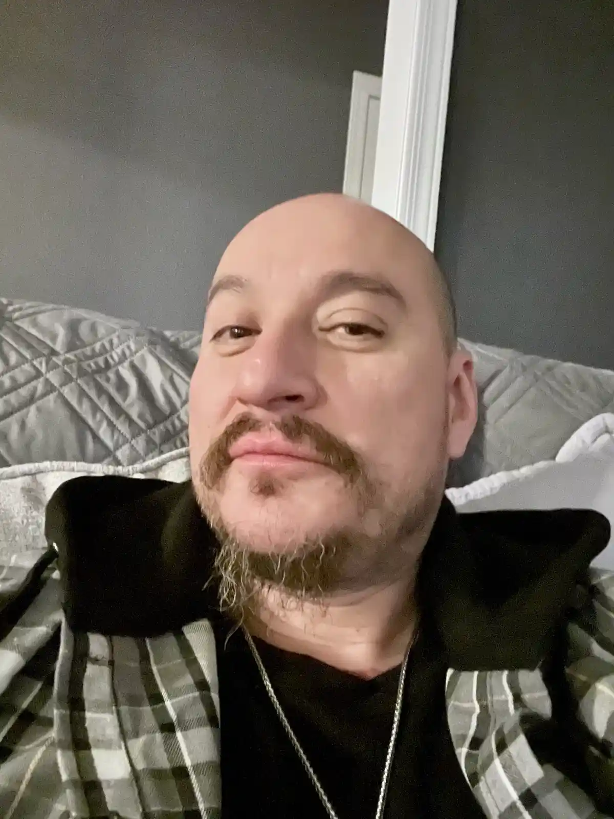 A bald man with a mustache and goatee, wearing a plaid shirt over a black shirt, lies on a bed with a gray quilted pillow…