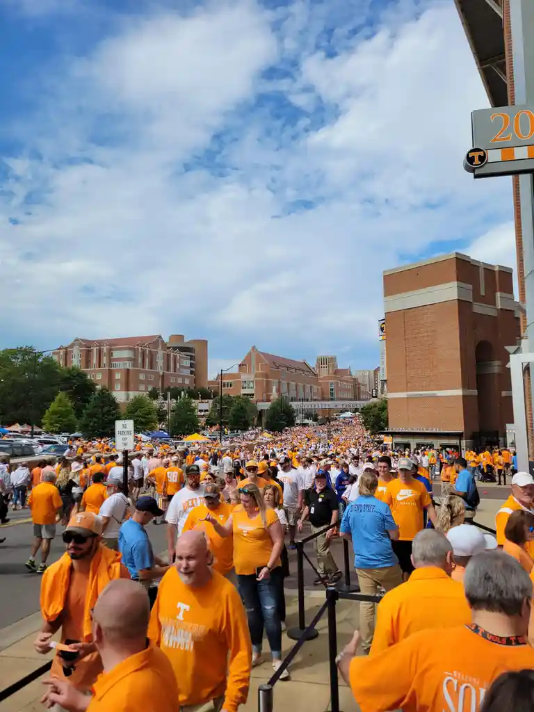 A large crowd of people, mostly wearing orange Tennessee Volunteers apparel, fills a street outside a stadium under a partly…