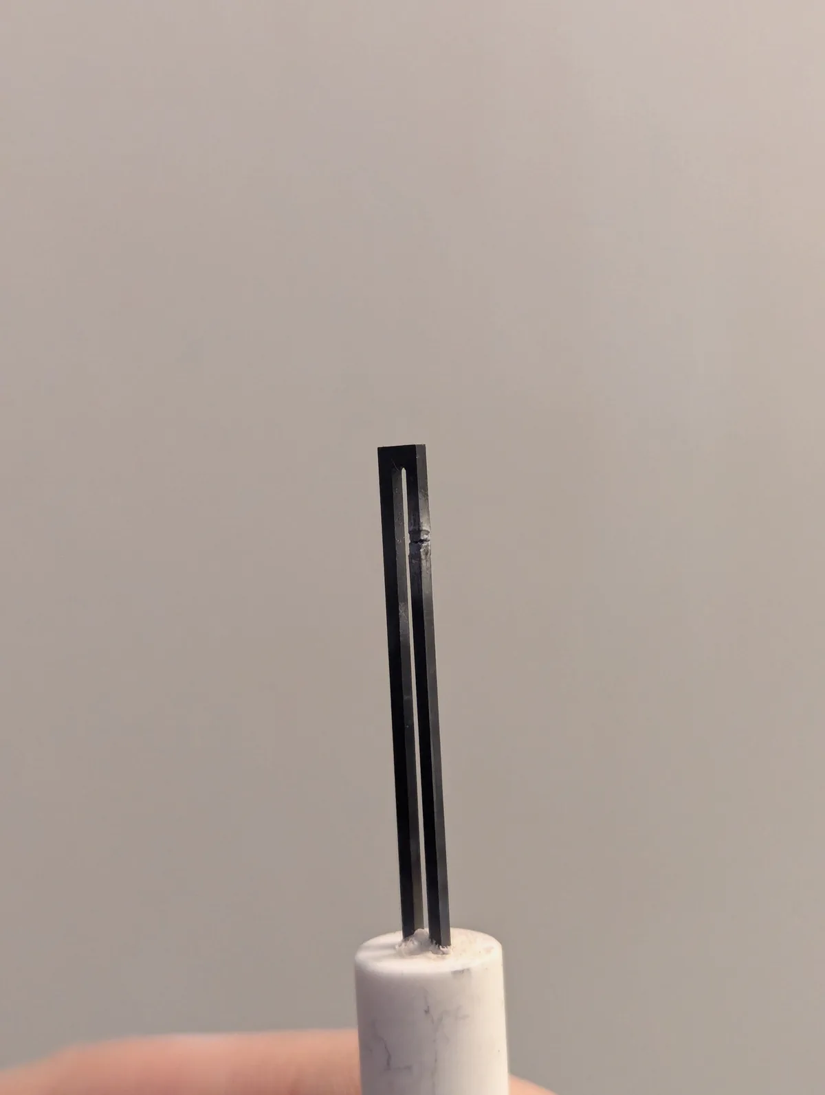 A close-up photo shows a black tuning fork standing upright in a white marble-like holder against a plain gray background. A…