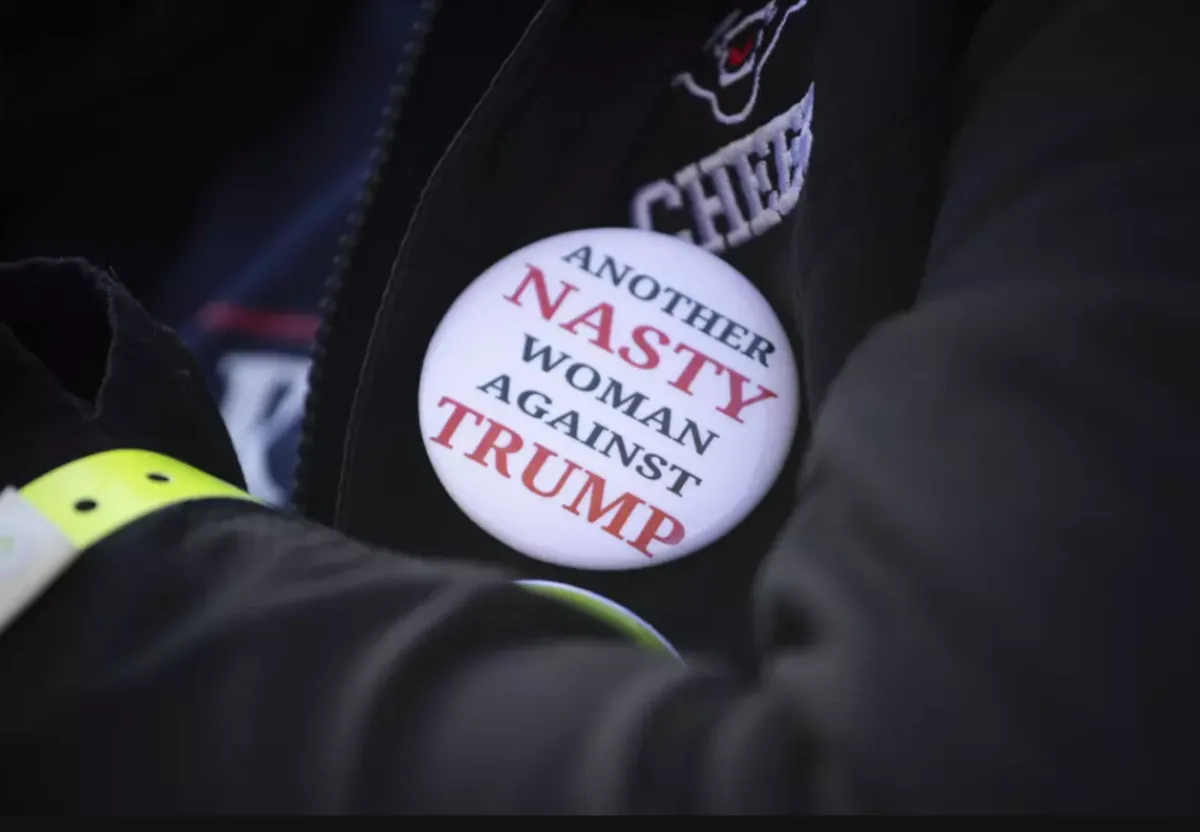 A close-up photo shows a white circular button pinned to a black jacket, with the text 'ANOTHER NASTY WOMAN AGAINST TRUMP'…