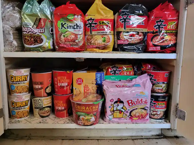 A pantry shelf is densely packed with various instant ramen products, including Nongshim Shin Ramyun (Black, Red, Gold)…