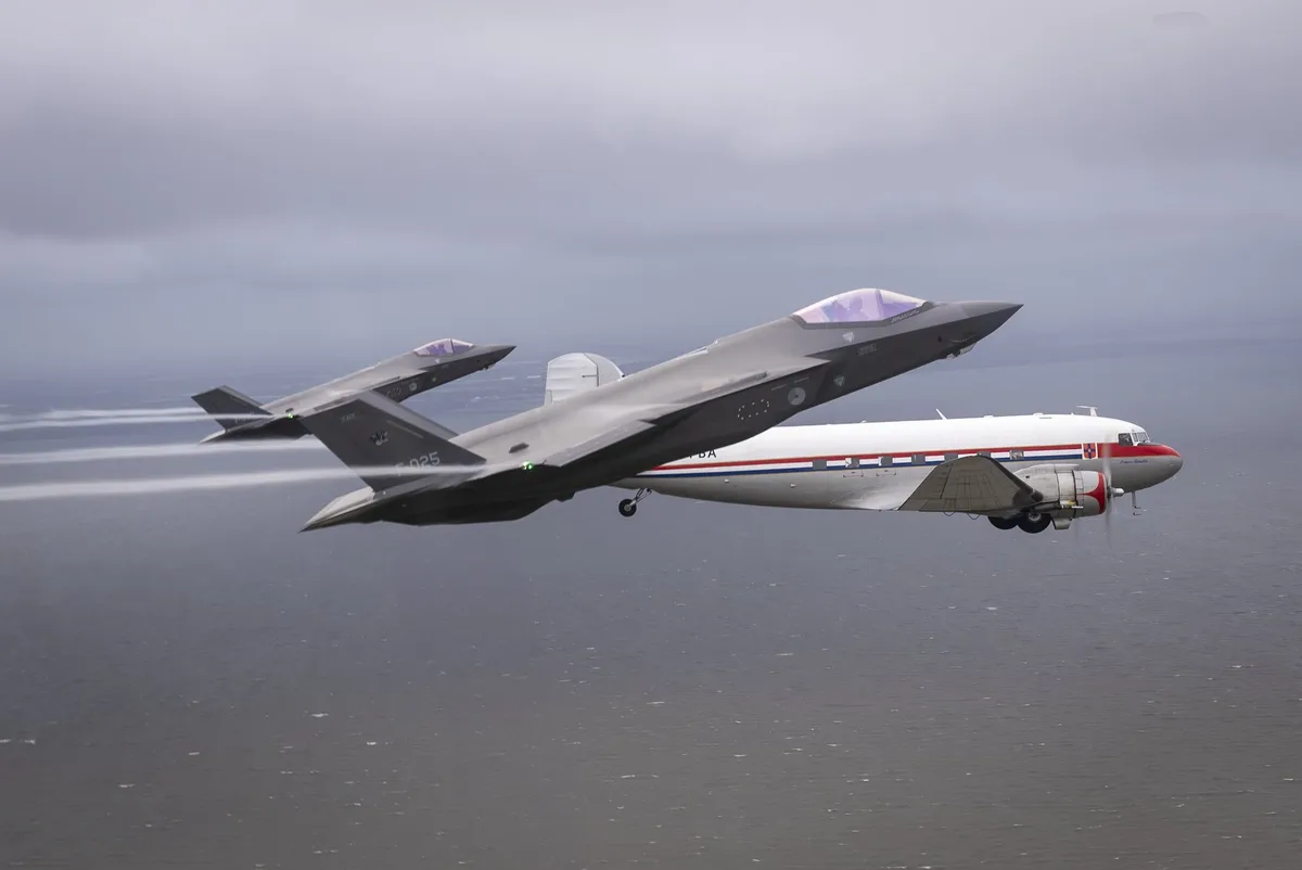 Two gray F-35 fighter jets fly in tight formation above a body of water, with a vintage white propeller plane flying below…