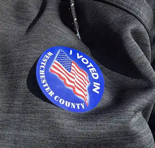 A close-up photo shows a blue circular sticker with an American flag design and the text 'I VOTED IN WESTCHESTER COUNTY'…