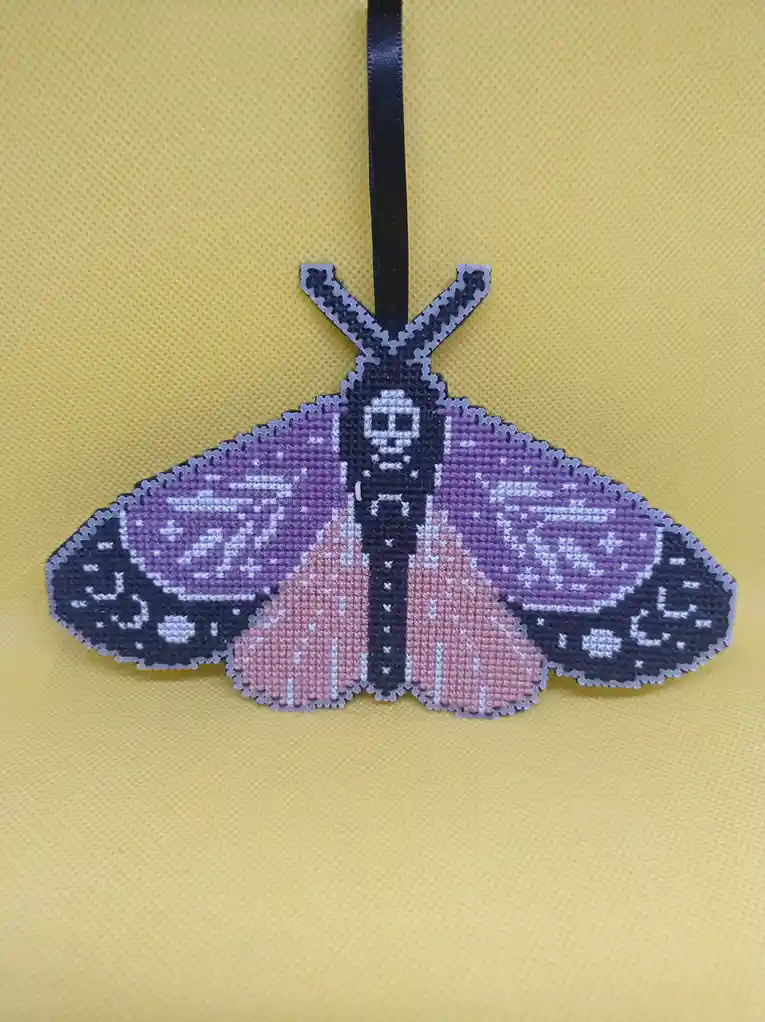 A cross-stitched moth ornament with purple, pink, and black wings hangs against a yellow textured background. The moth's…