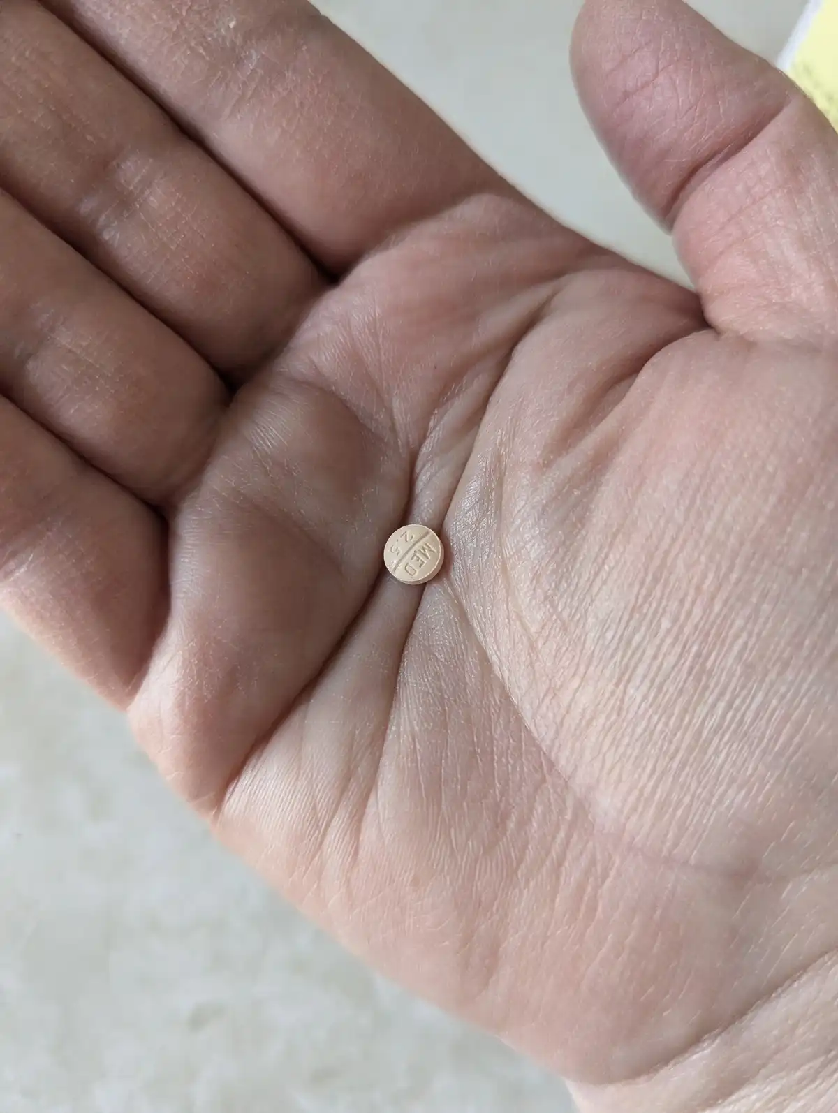 A close-up photo shows a person's open palm holding a small, round, beige pill with 'MED 2.5' embossed on it. The hand's…