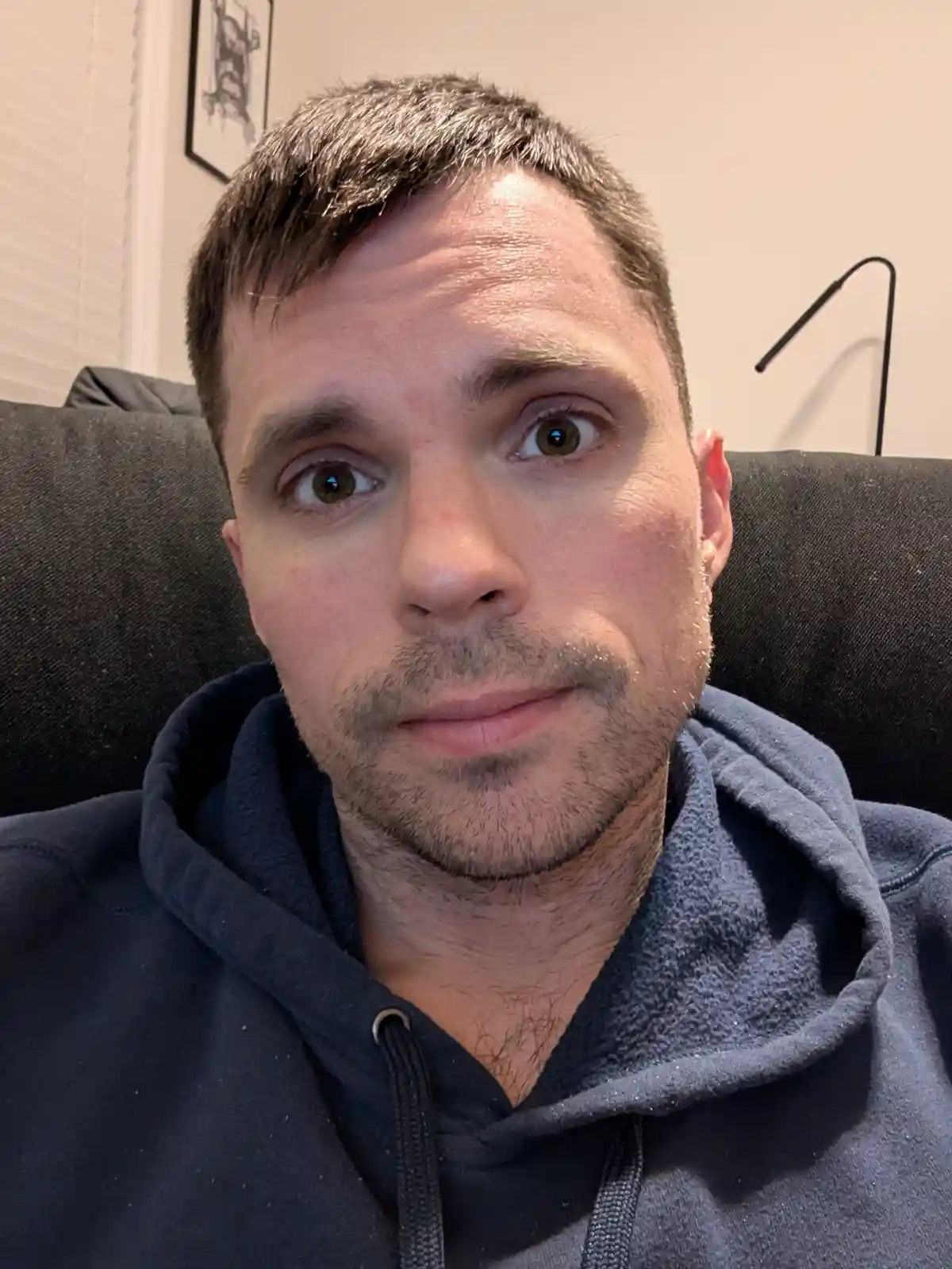 A man with short brown hair and a light beard, wearing a dark blue hoodie, sits on a dark couch and stares directly at the…