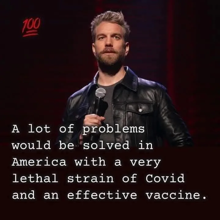 A man with short blond hair and a beard, wearing a black leather jacket over a gray shirt, stands on a dimly lit stage…