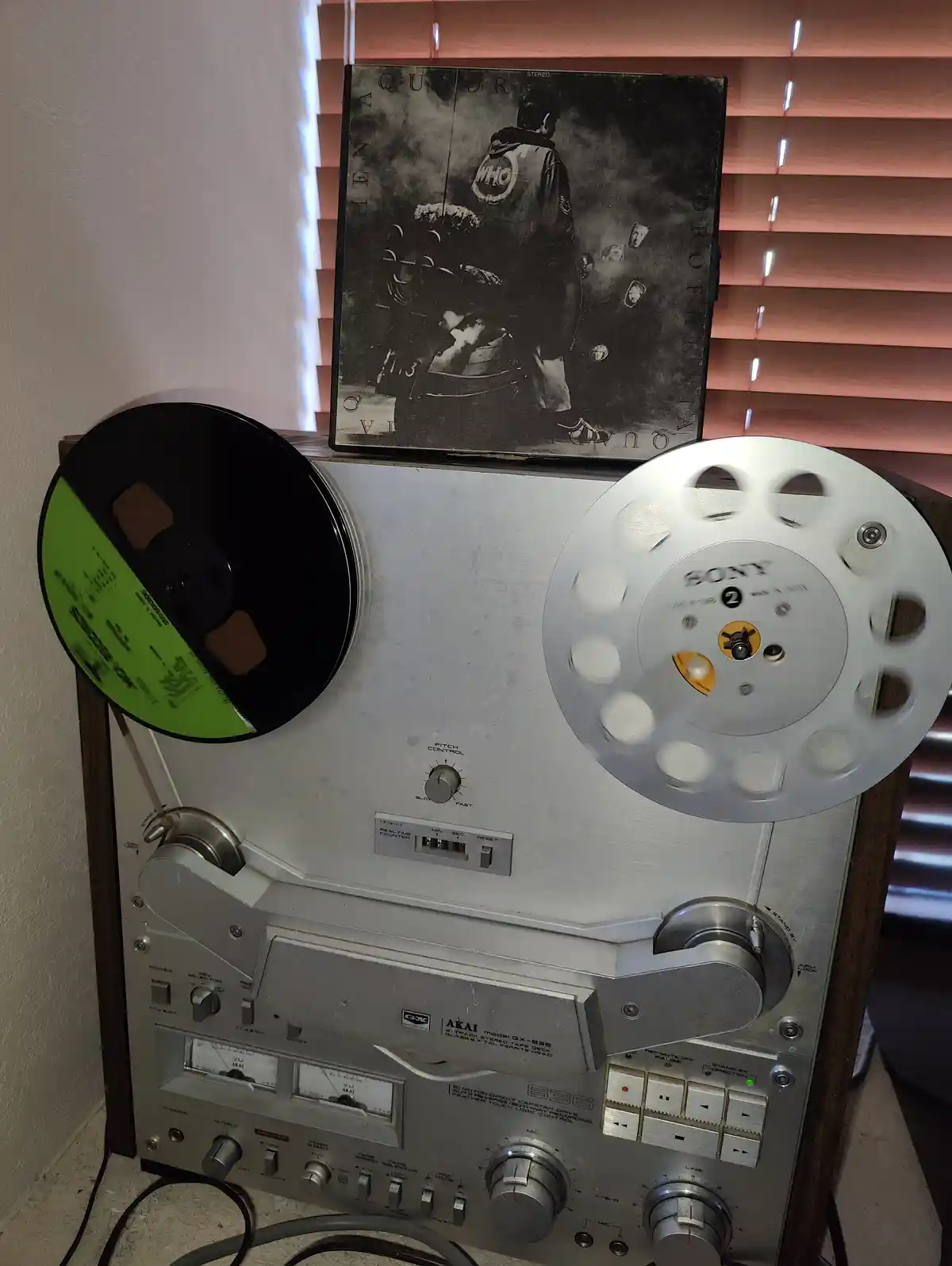 A vintage Akai reel-to-reel tape deck with a green-labeled tape reel on the left and a white Sony reel on the right, with a…