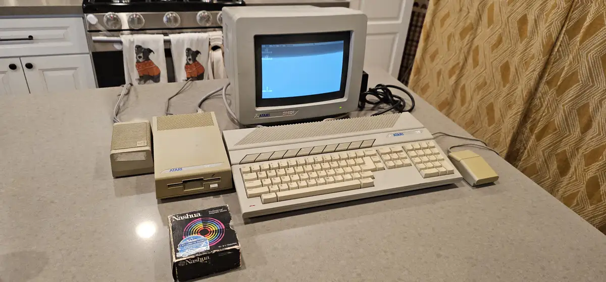 An Atari ST computer with monitor, keyboard, mouse, and floppy disk drive sits on a modern kitchen countertop, next to a box…