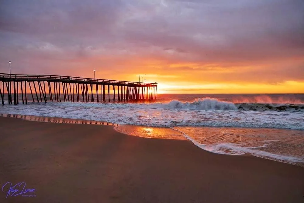 A serene beach sunset photo featuring a long wooden pier extending into the ocean, with waves crashing near the shore and a…