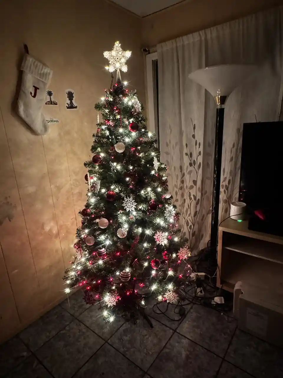 A decorated christmas tree with red and white ornaments and lights stands in a dimly lit room next to a tv stand and a floor…