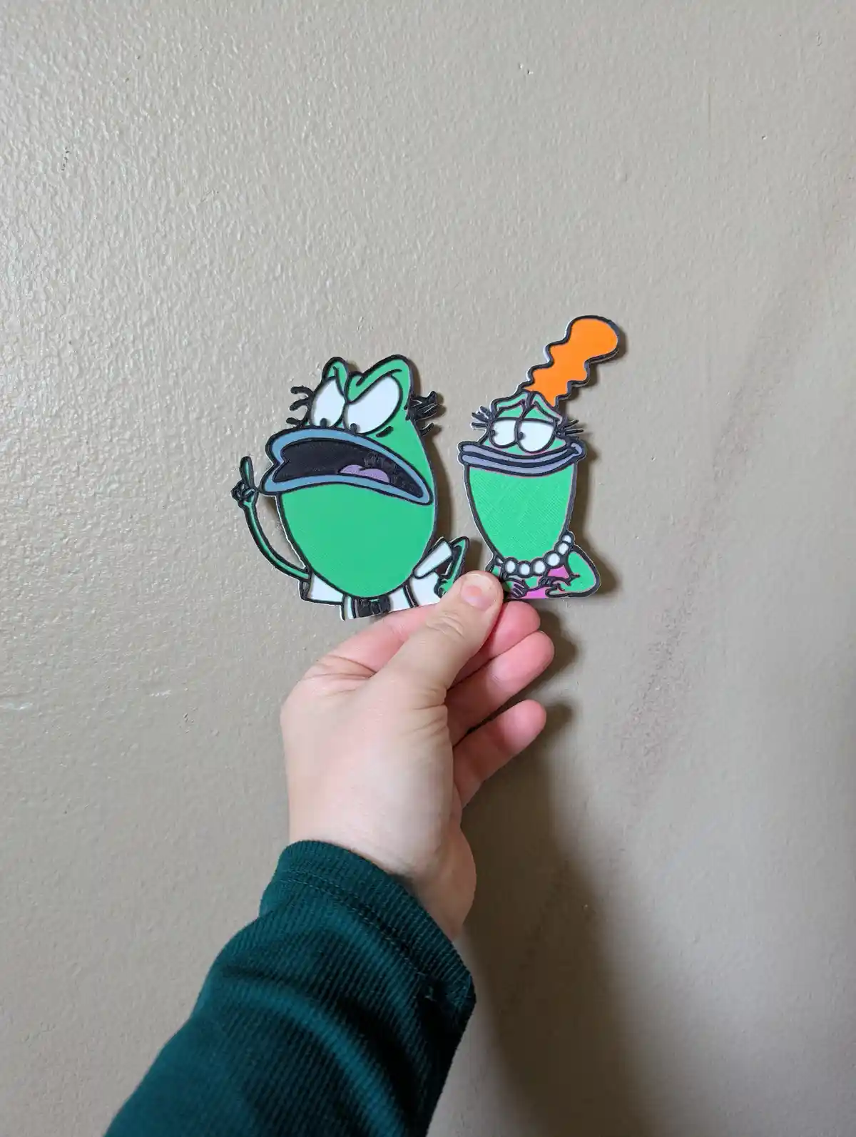 A hand holds two green cartoon frog cutouts against a beige wall. The frog on the left has an angry expression and is…
