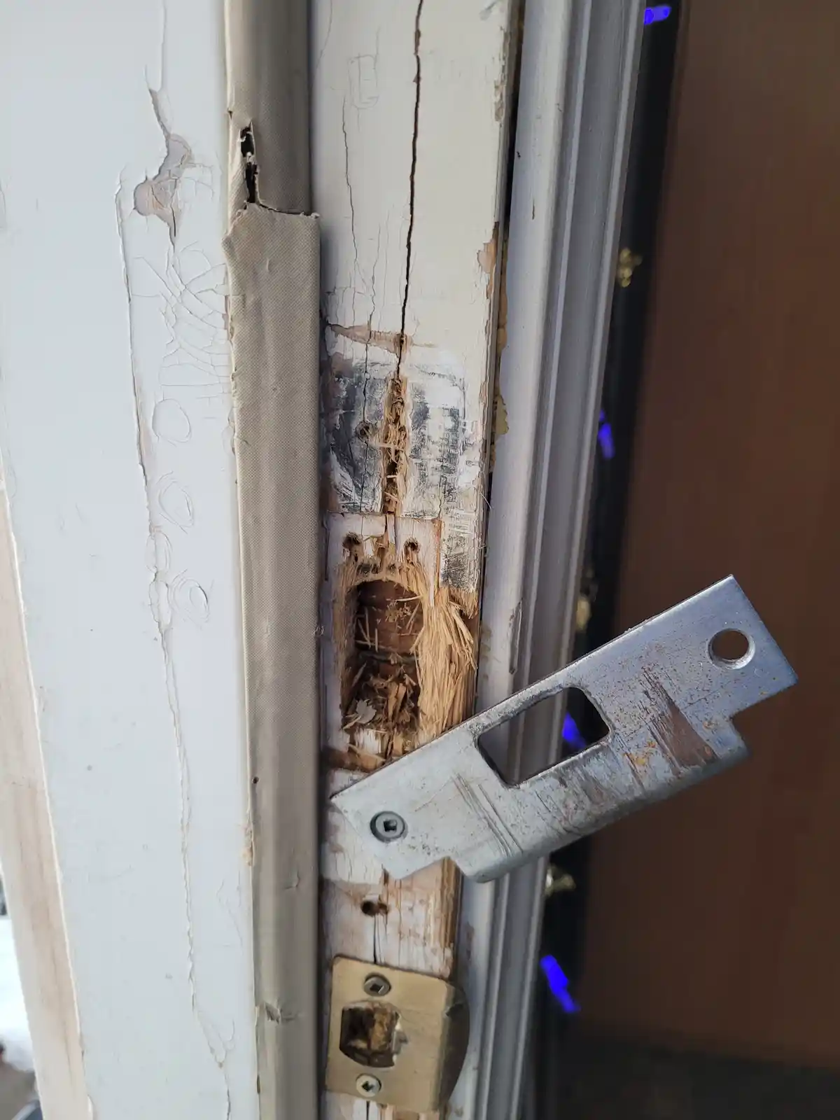 A close-up photo shows a severely damaged wooden door frame with peeling white paint, splintered wood, and a large hole…