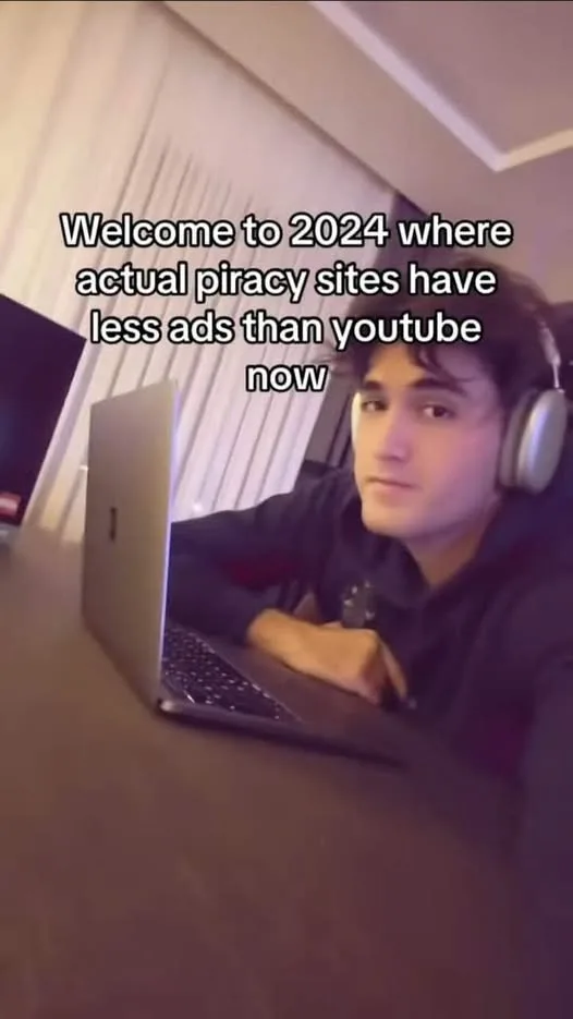 A young man wearing headphones sits at a desk with a laptop open in front of him, looking directly at the camera with a…