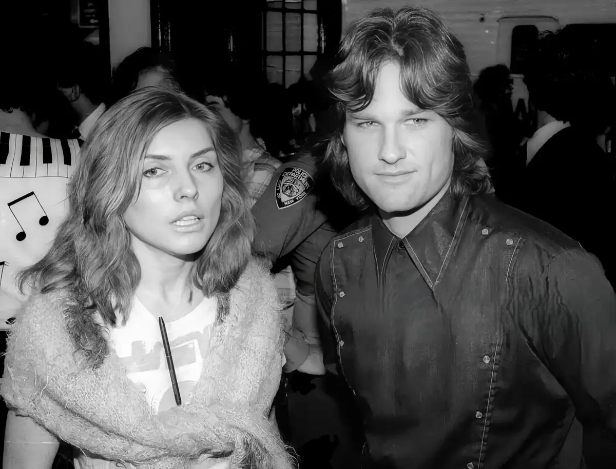 A black-and-white photo shows a woman with a skeptical, unimpressed expression standing next to a man with a calm, slightly…