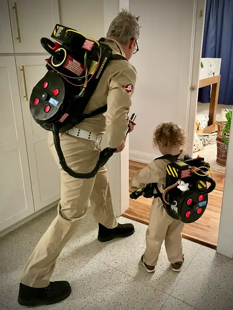 An adult and a small child, both dressed in Ghostbusters costumes complete with proton packs and ghostbusting gear, walk…