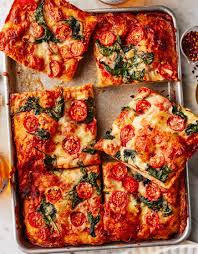 A close-up photo of a rectangular pizza on a metal baking sheet, cut into several square slices. The pizza has a thick…