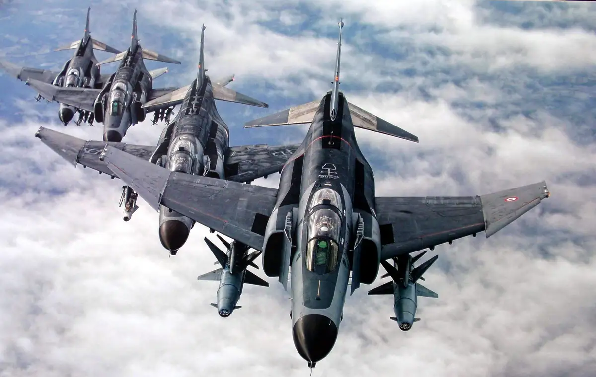 Four fighter jets fly in tight formation above a layer of clouds, with the lead aircraft prominently displayed. The jets are…