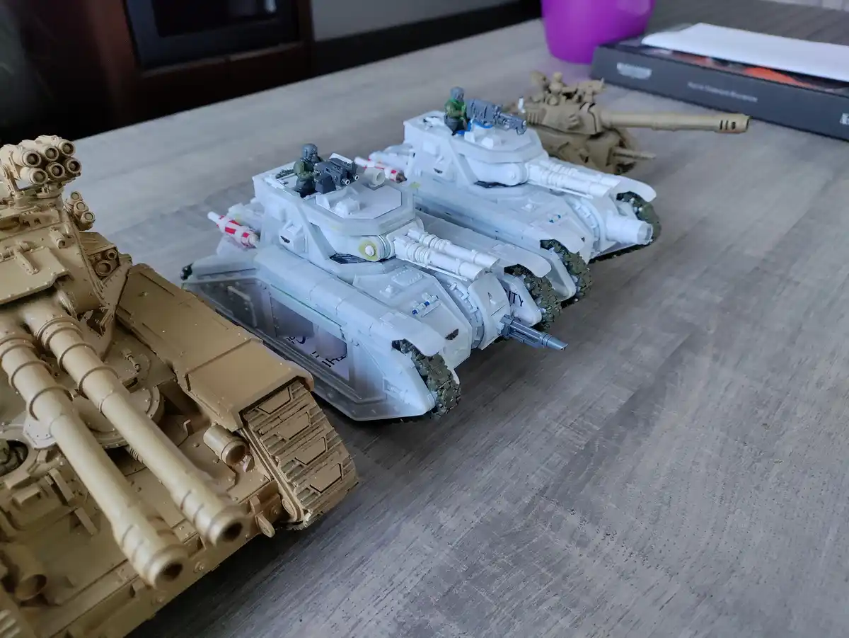 Three model tanks are lined up on a wooden table, two white and one tan, with miniature soldiers on top. The white tanks…