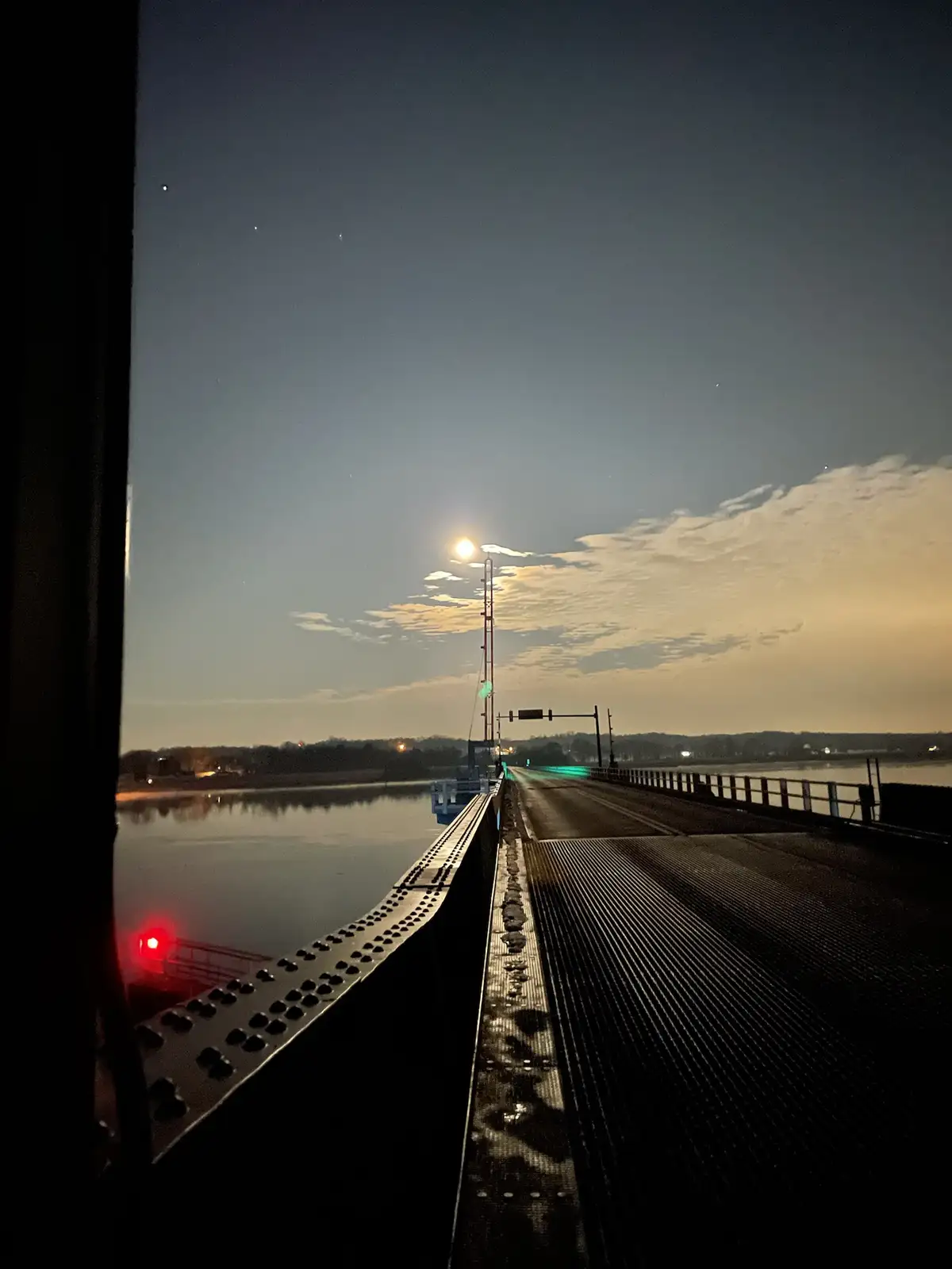 A nighttime photo taken from a vehicle on a bridge over water, showing a moonlit sky with scattered clouds, a red light on…