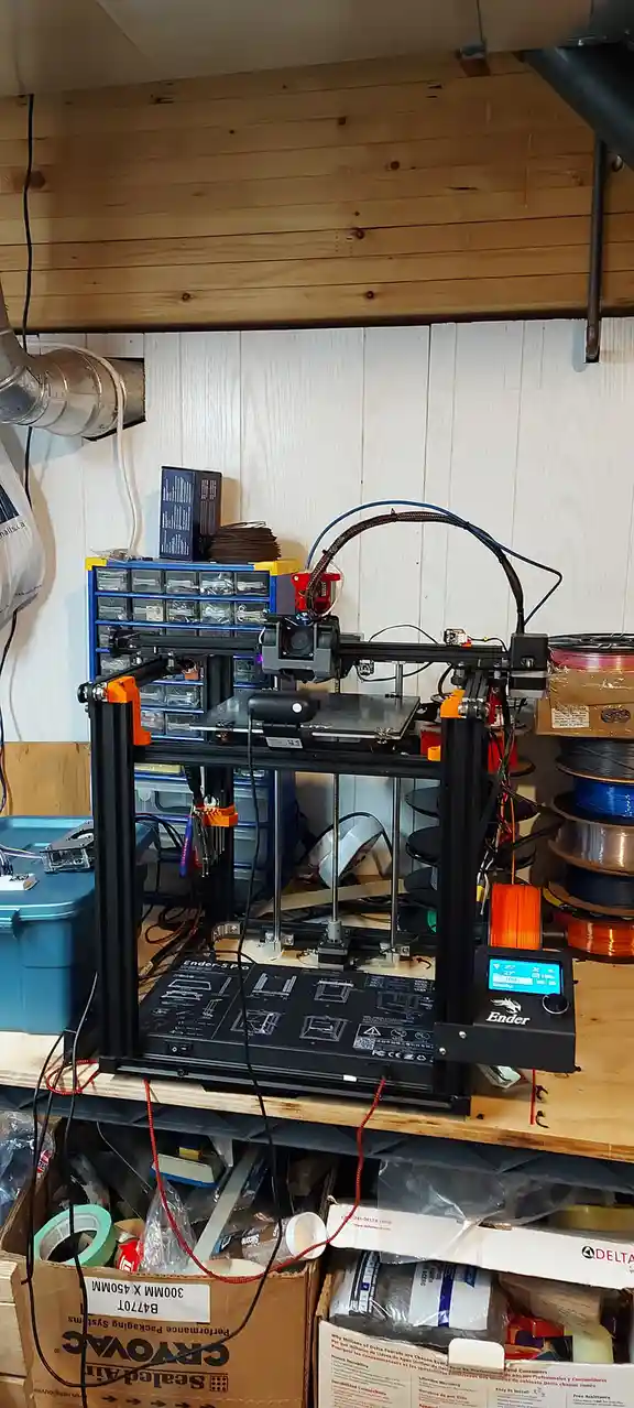 A black Ender-5 Pro 3D printer sits on a wooden shelf in a cluttered workshop, surrounded by spools of filament, toolboxes…