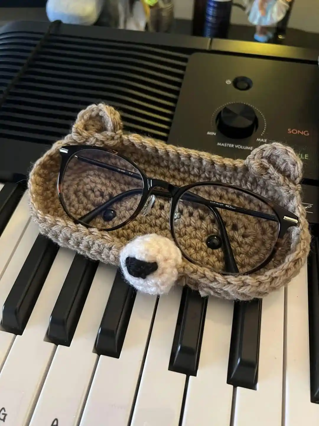 A pair of black-rimmed glasses rests inside a handmade, brown crocheted bear-shaped case with ears and a nose, placed on the…