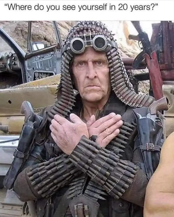 A man in a post-apocalyptic setting, wearing goggles and a hood made of ammunition belts, has his hands clasped over his…
