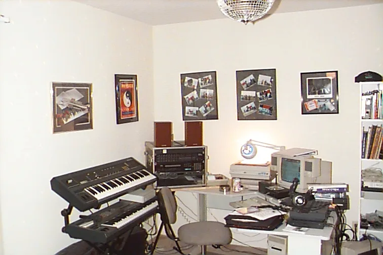 a cluttered home studio room with two keyboards, a computer, speakers, and framed pictures on the walls, with the text 'this…
