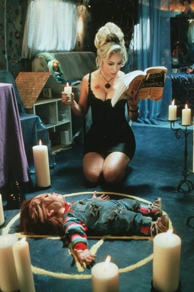 A woman in a black dress kneels in a circle of lit candles, holding a candle and reading a book titled 'Voodoo for Dummies'…
