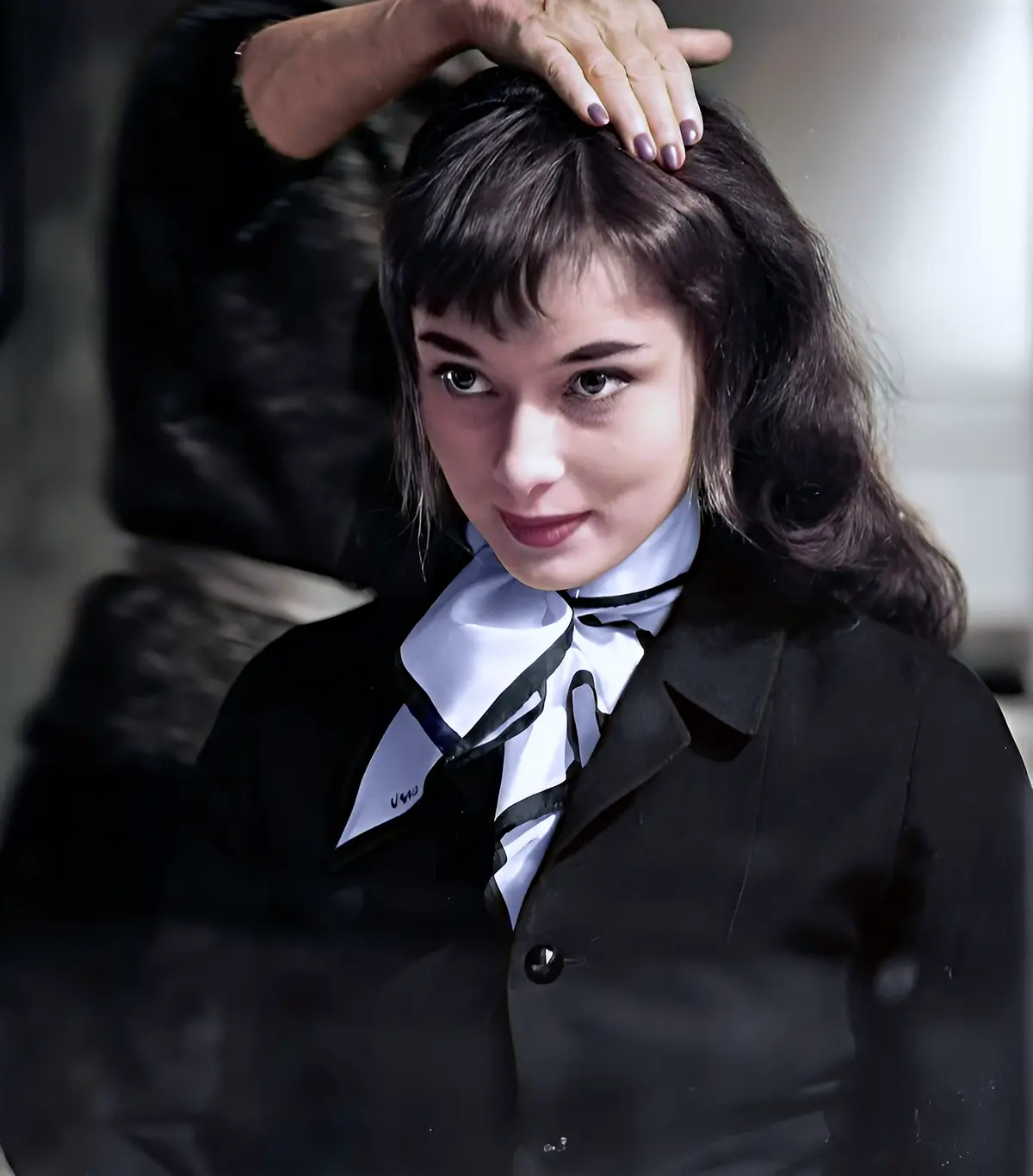 A woman in a black blazer and white shirt with a black bow tie looks directly at the camera with a slight smirk while a hand…