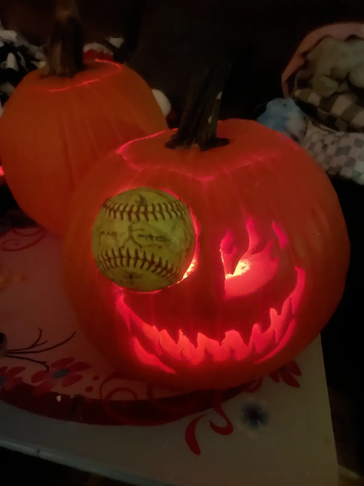 A carved Halloween pumpkin with a glowing red interior and a jagged, menacing face, has a yellow softball inserted into one…