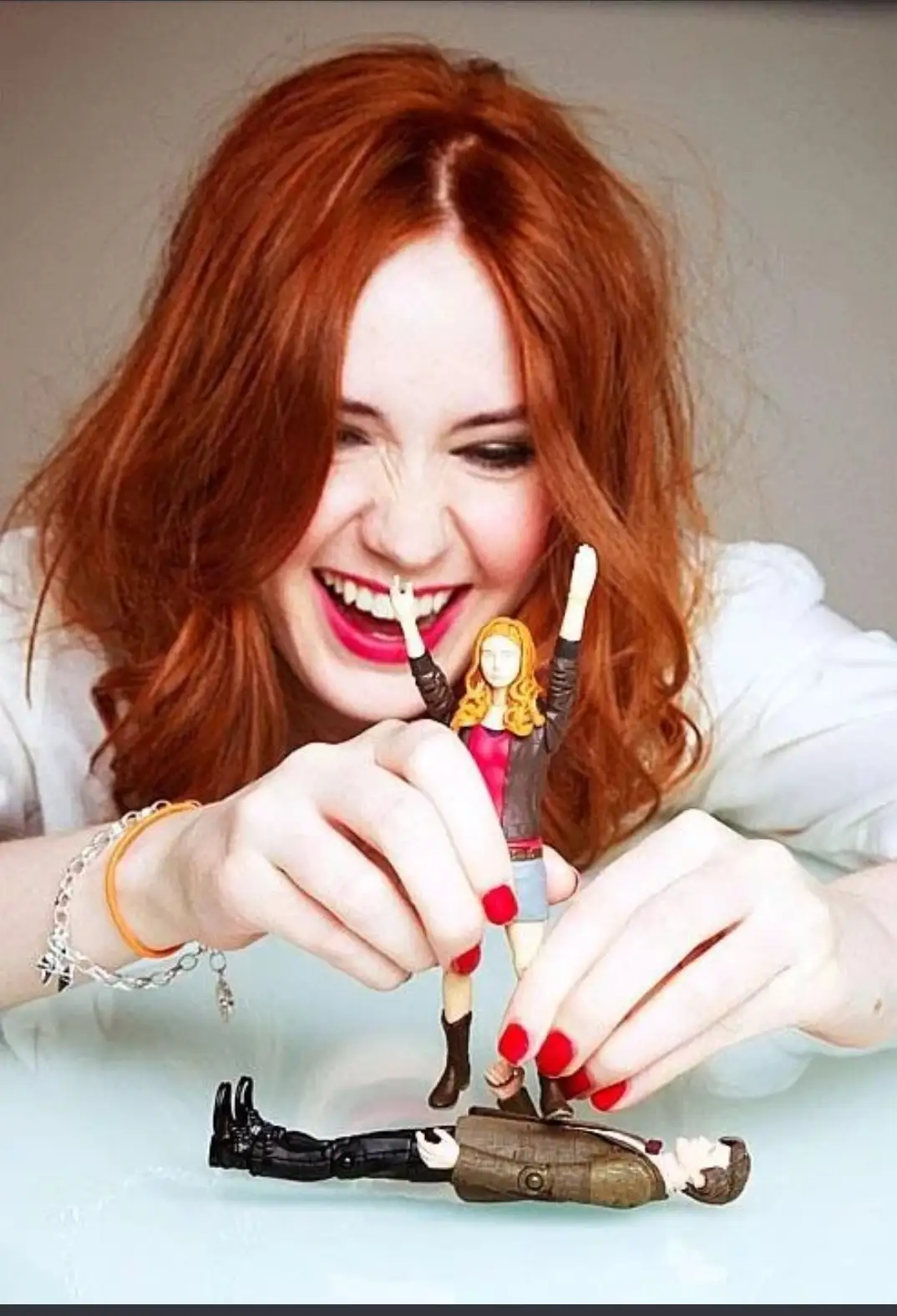 A woman with red hair and bright red fingernails holds a small figurine of a woman with blonde hair, arms raised in triumph…