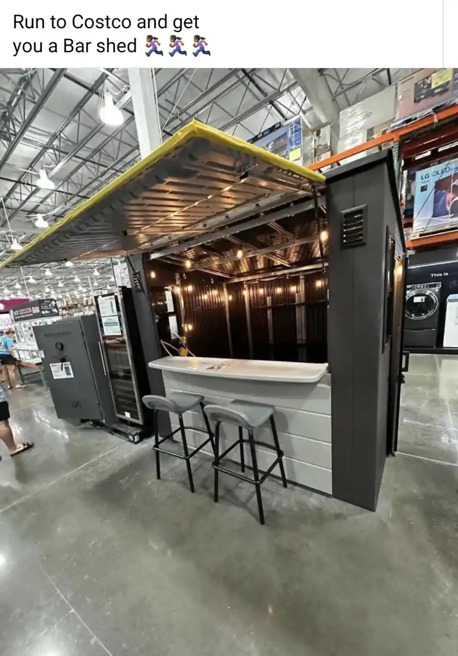 A photo of a small, freestanding bar structure with a white countertop, two grey bar stools, and a dark interior with string…