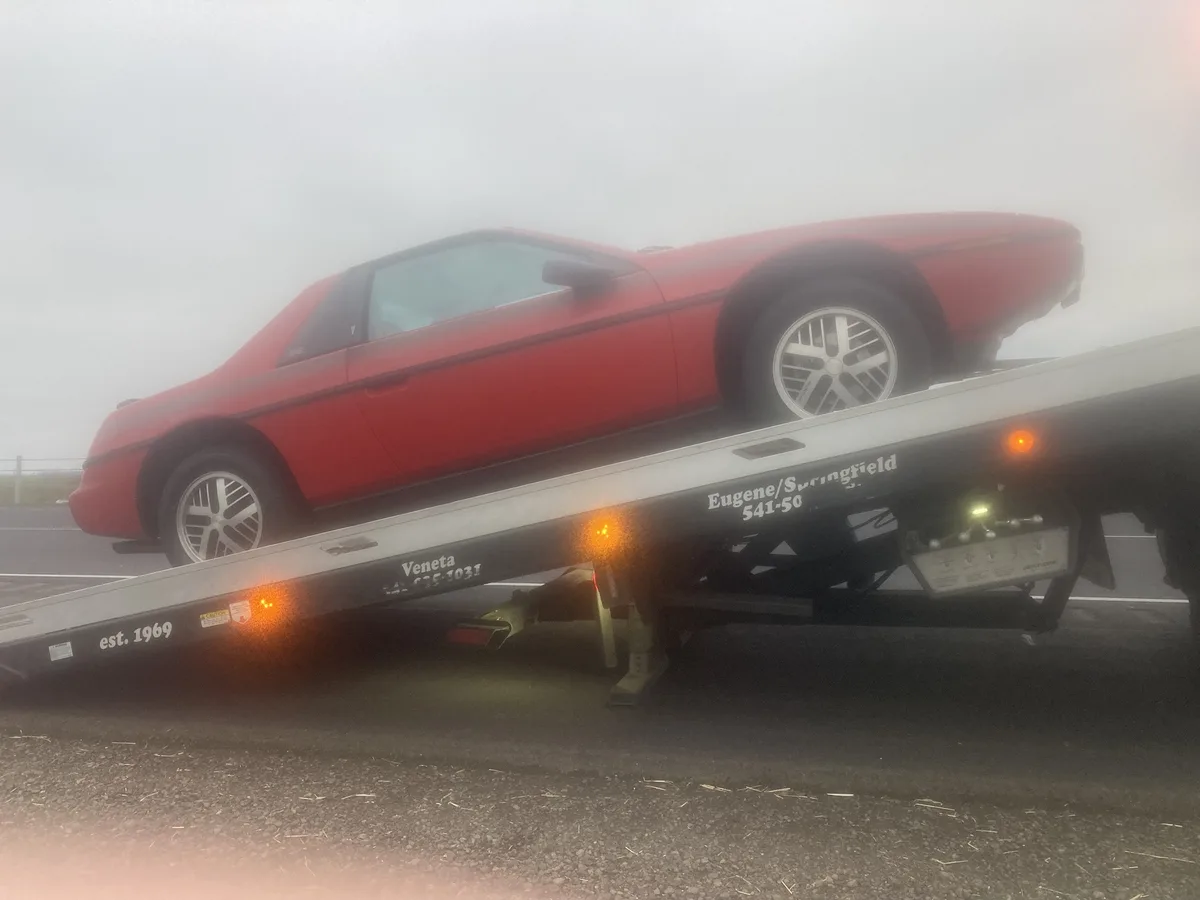 A red sports car is loaded onto a flatbed tow truck in thick fog. The truck has text reading 'Veneta est. 1969' and…