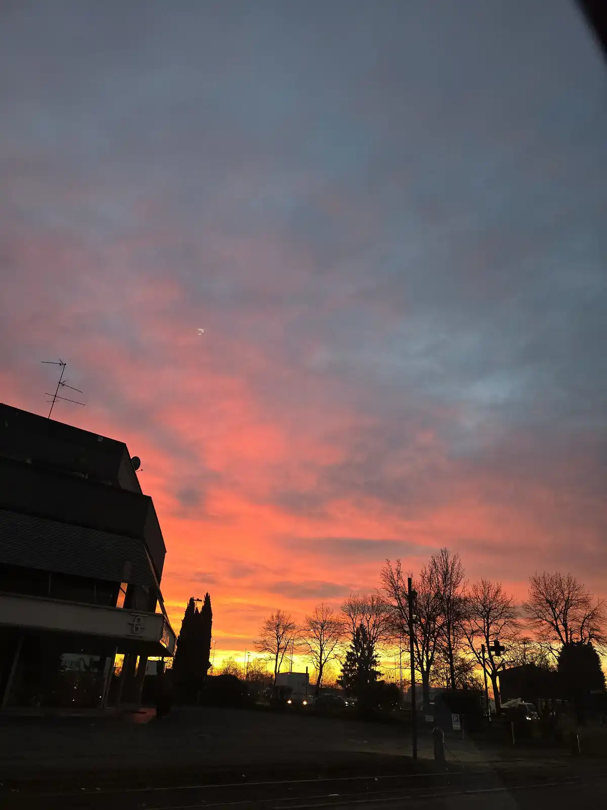 A photograph captures a vibrant sunset with fiery orange and pink clouds against a dusky blue sky, silhouetting bare trees…