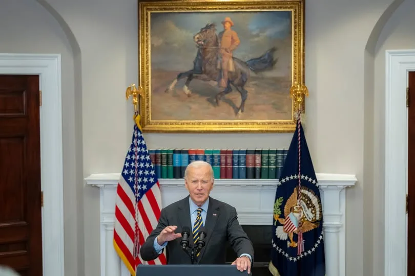 a man in a suit stands at a podium in an official room with american flags and a fireplace. Above him hangs a large painting…