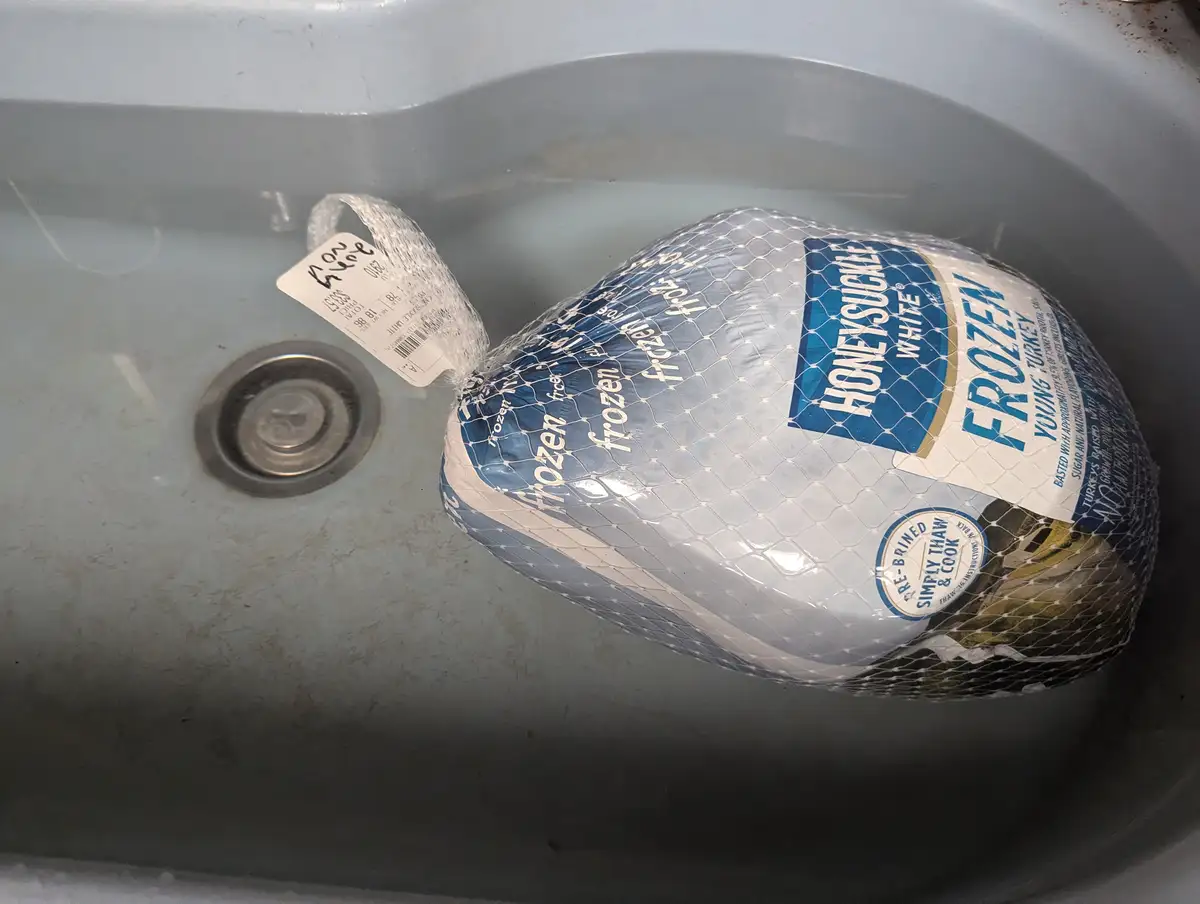 A frozen Honeysuckle White young turkey, still in its plastic netting and branded packaging, is sitting in a sink filled…