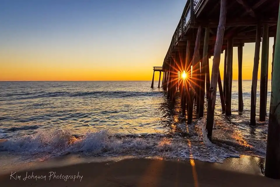 A serene sunrise photo showing the sun rising directly behind the wooden pilings of a pier, casting a bright starburst…