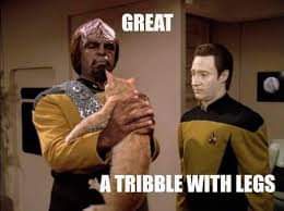 A Star Trek character in a yellow uniform holds a small, furry creature while another character looks on. White text…