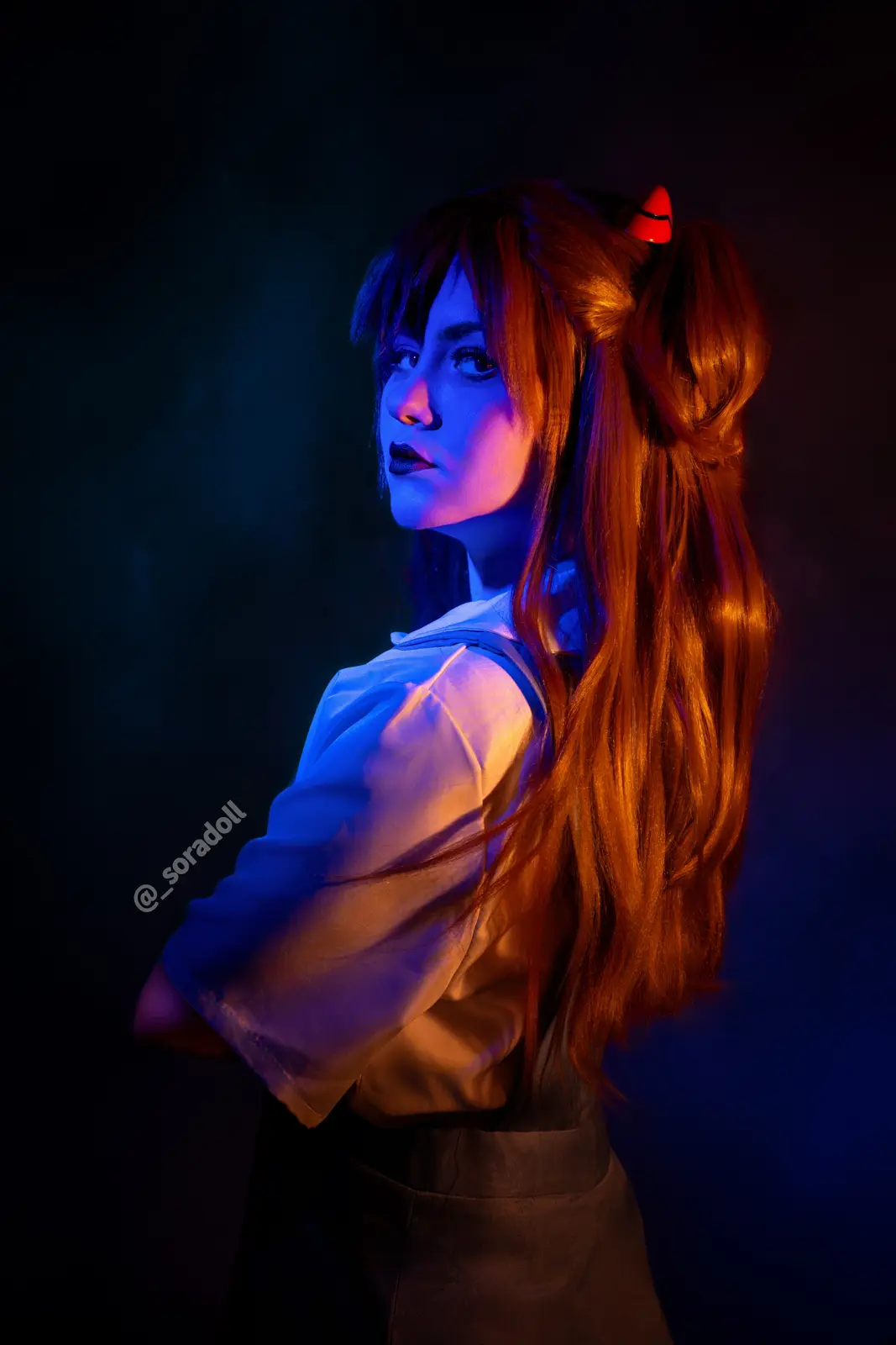A person in cosplay with long red hair styled in a high ponytail, featuring a small red devil horn accessory. They wear a…