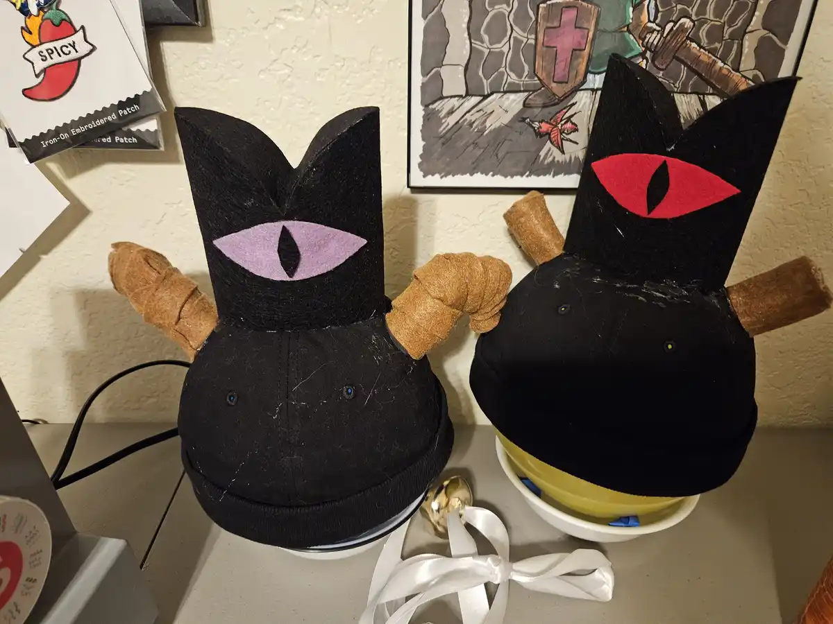 Two black baseball caps are modified into cat-like figures with felt ears, large stylized eyes (one purple, one red), and…