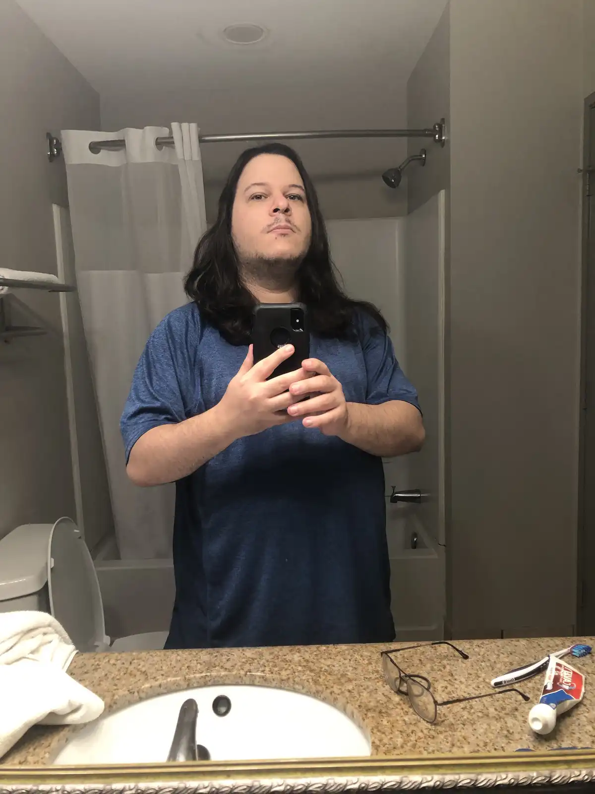 A man with long dark hair and a blue t-shirt takes a mirror selfie in a bathroom, holding a black phone. Behind him is a…