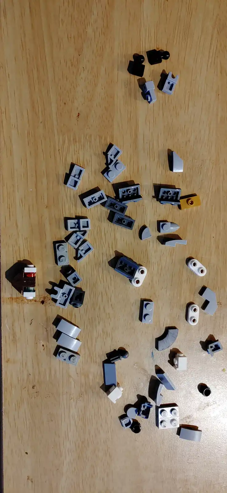 A top-down photo shows numerous disorganized Lego pieces of various shapes and colors scattered across a light wooden…