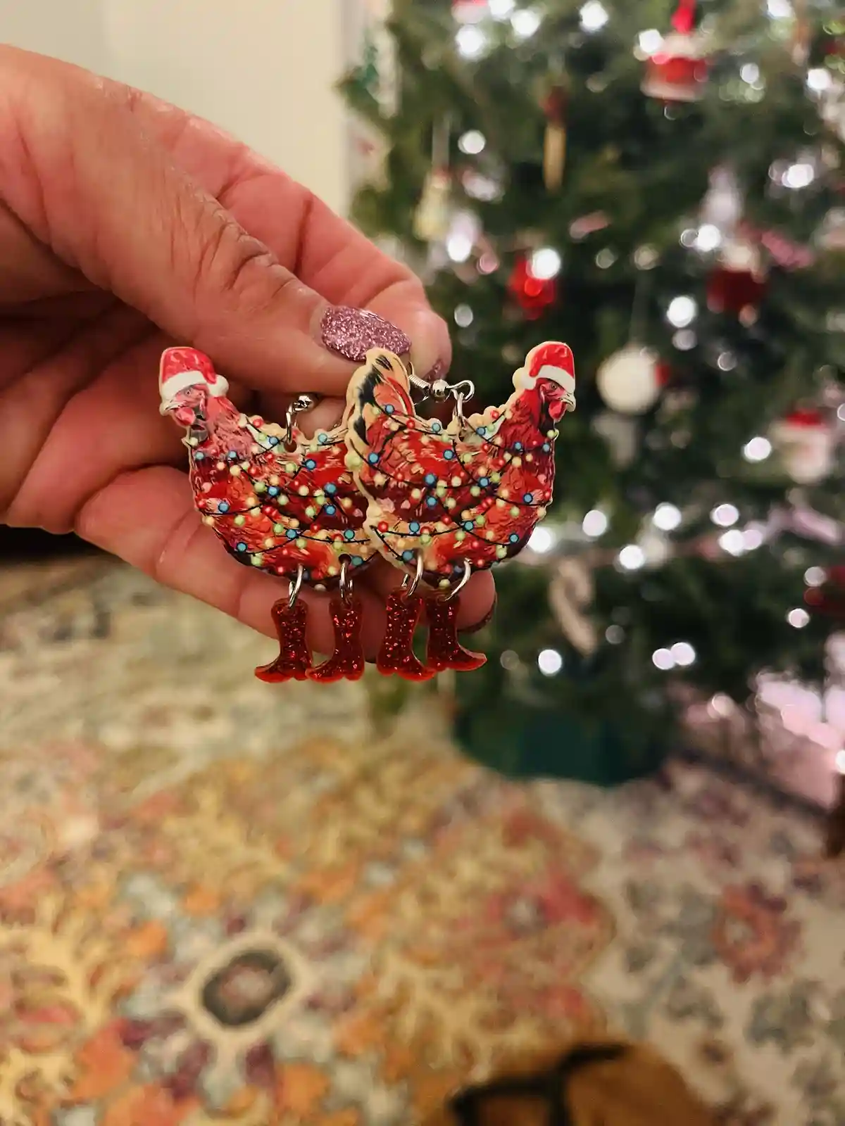 A hand with glittery pink nails holds a pair of earrings shaped like red chickens wearing Santa hats and decorated with…