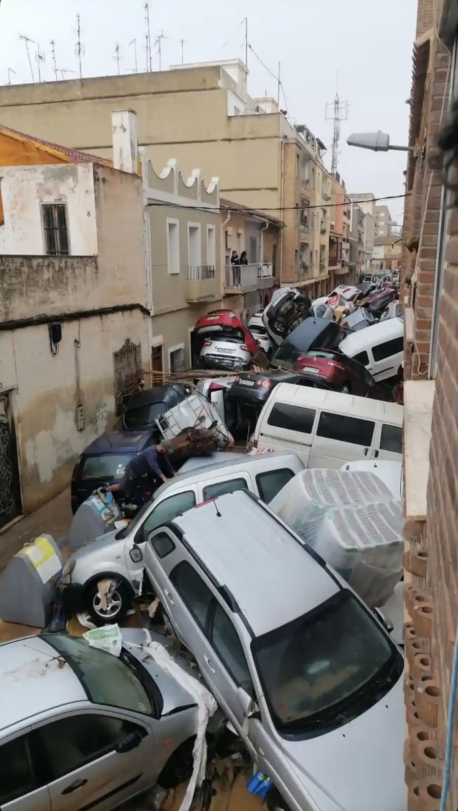 A narrow urban street is completely blocked by a chaotic pile of crushed, overturned, and damaged cars, with debris and…