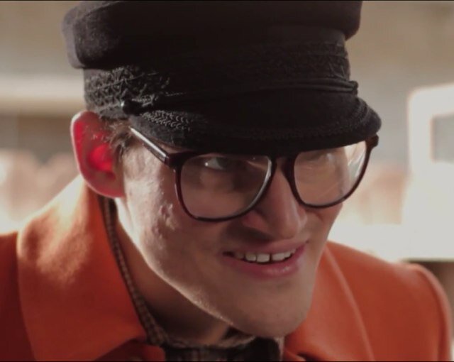 a close-up of a man wearing a black cap, thick-rimmed glasses, and an orange jacket, smiling smugly while looking slightly…